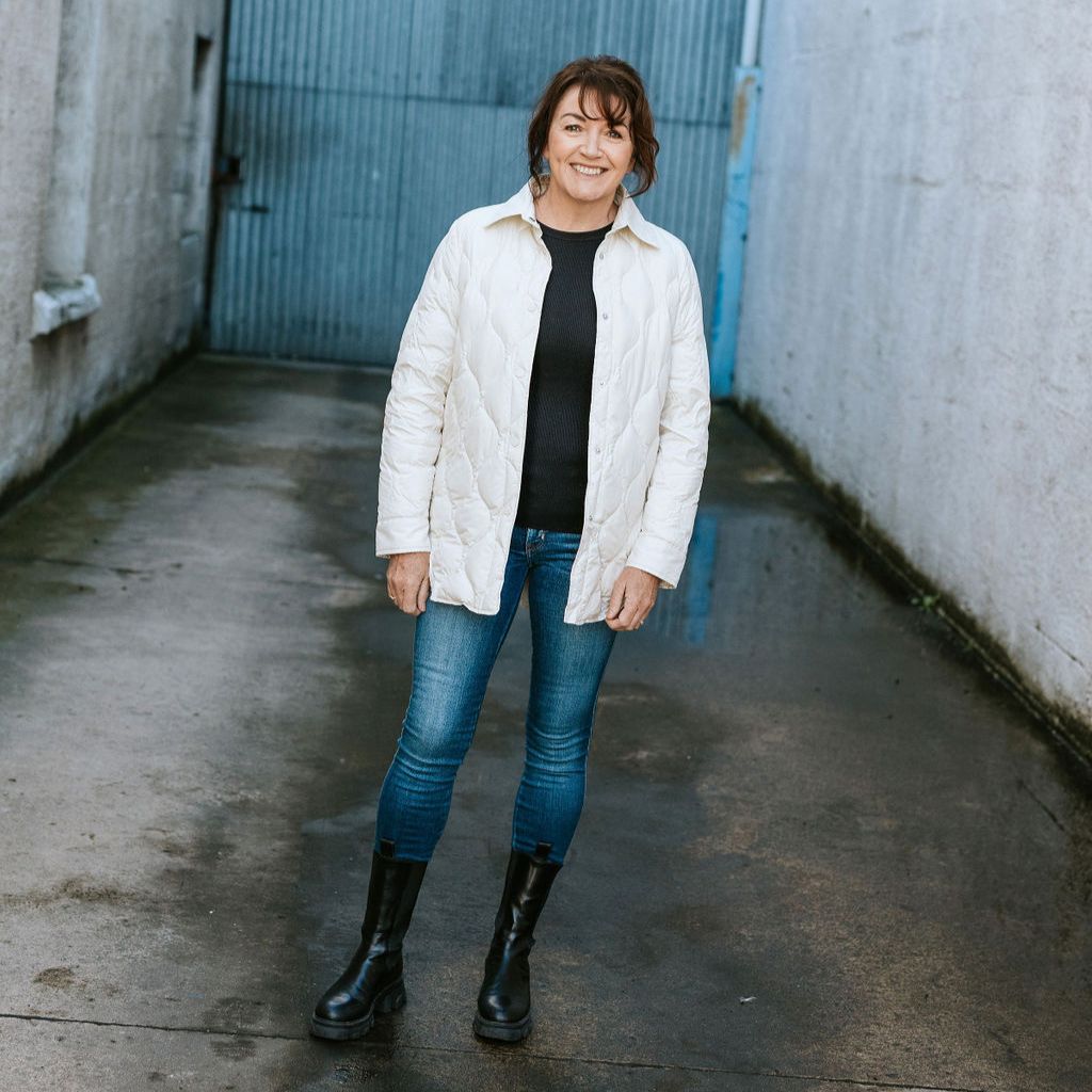 Woman wearing a white jacket, black top, blue jeans, and black boots standing in an alleyway.