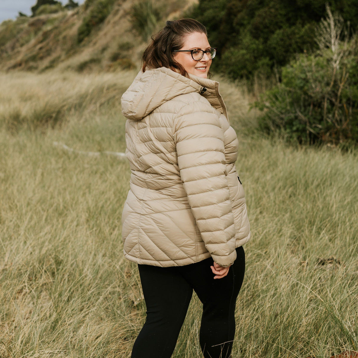 Woman wearing a Moke beige hooded puffer jacket standing in a grassy field.