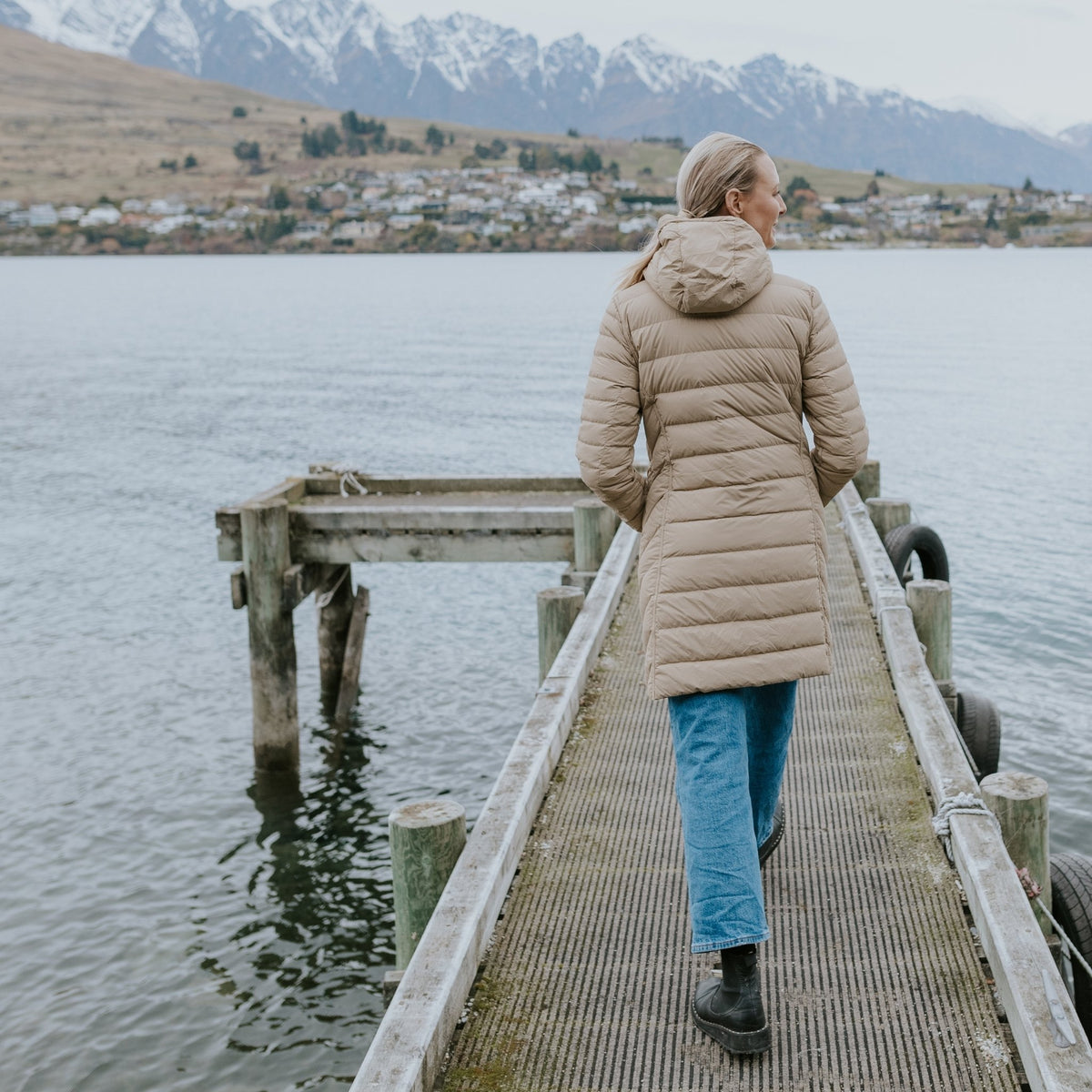 Woman wearing a beige Moke puffer jacket walking on a wooden pier by a lake and mountains.