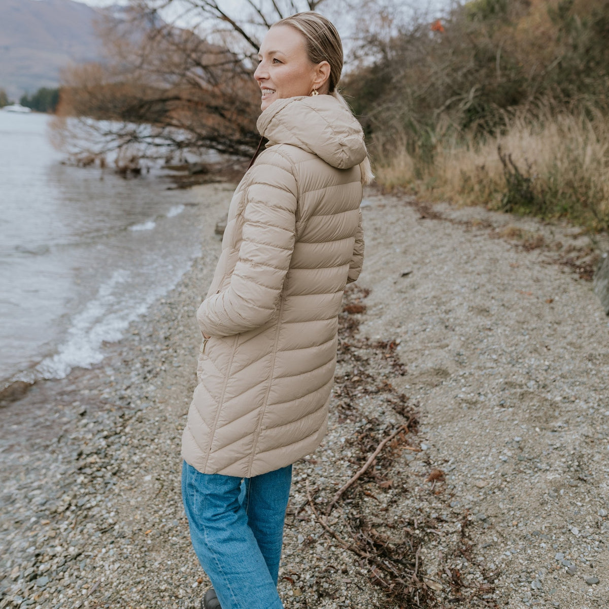 Woman wearing a Moke beige puffer jacket and blue jeans standing by a lake shore.