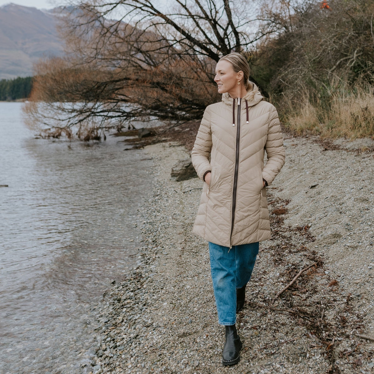 Woman wearing a Moke beige quilted puffer jacket and blue jeans walking on a rocky lakeshore.