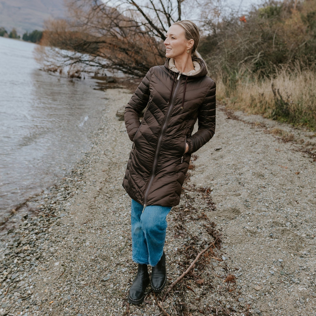 Woman wearing a Moke brown puffer jacket and blue jeans on a stony lakeshore.