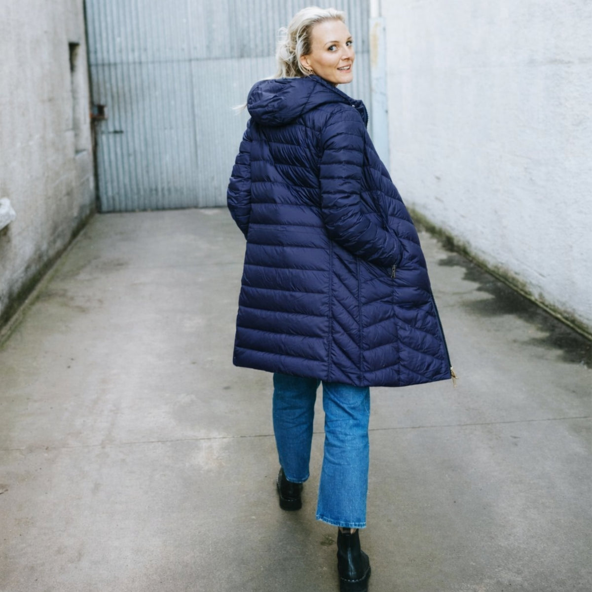 Woman wearing a navy blue Moke long puffer jacket and blue jeans walking in an alley.