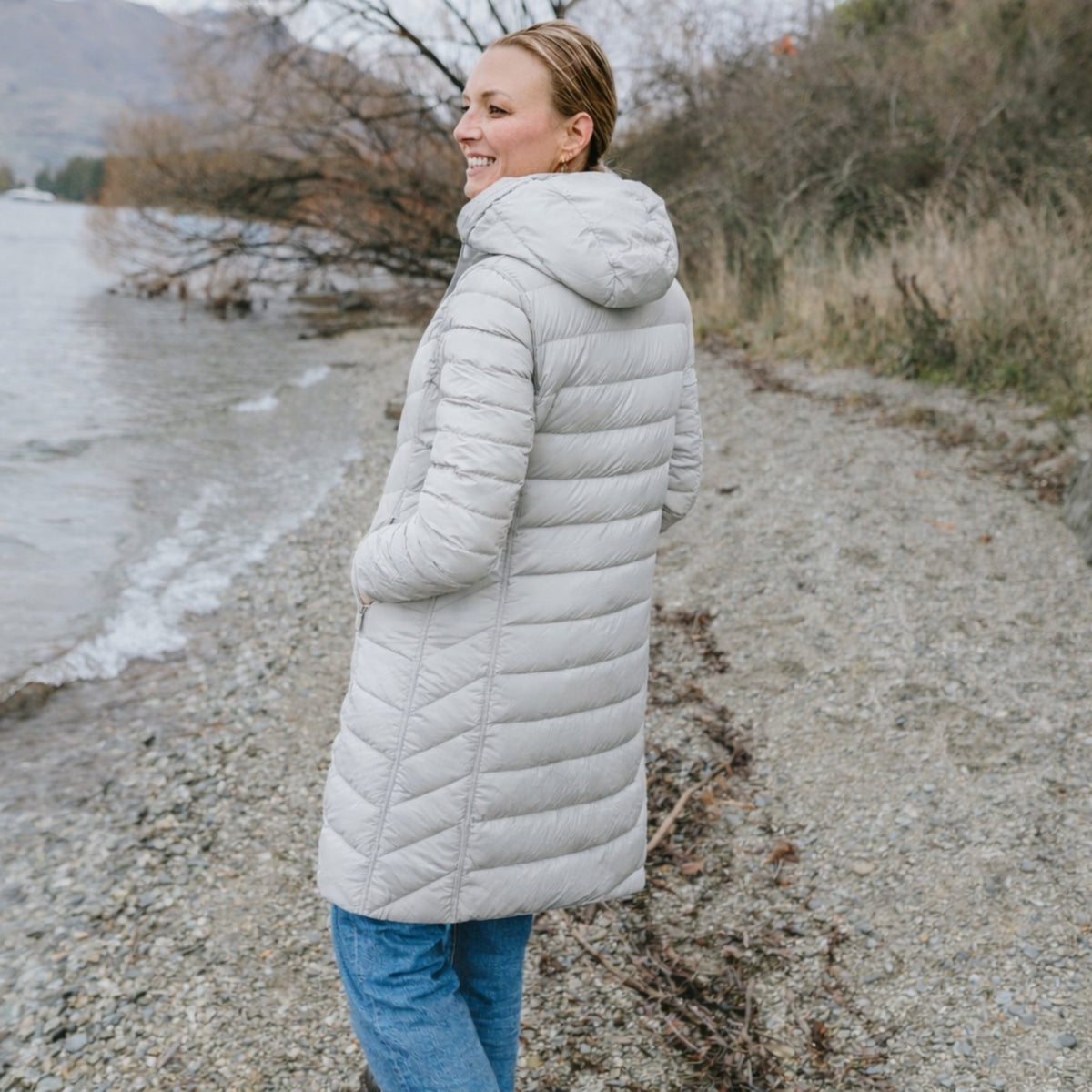 Woman wearing a Moke long light grey puffer jacket with hood by a lake shore.
