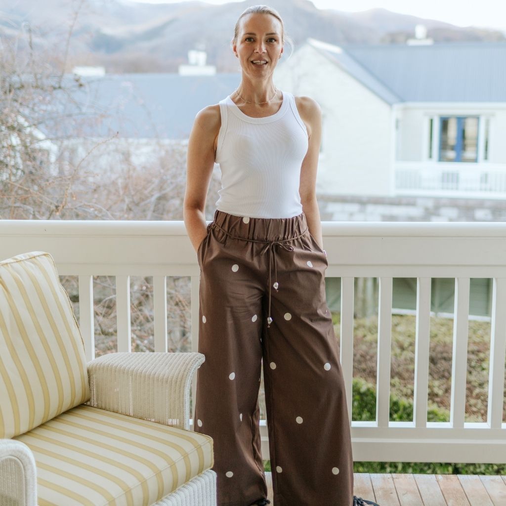 Woman wearing a white ribbed tank top and brown polka dot Moke trousers on a balcony.