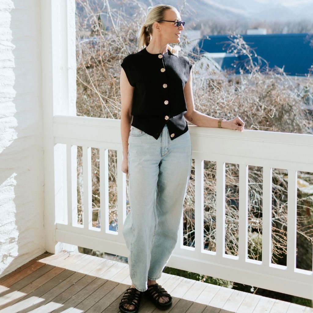 Woman wearing a black Moke button-up knit vest and light wash jeans on a white balcony.