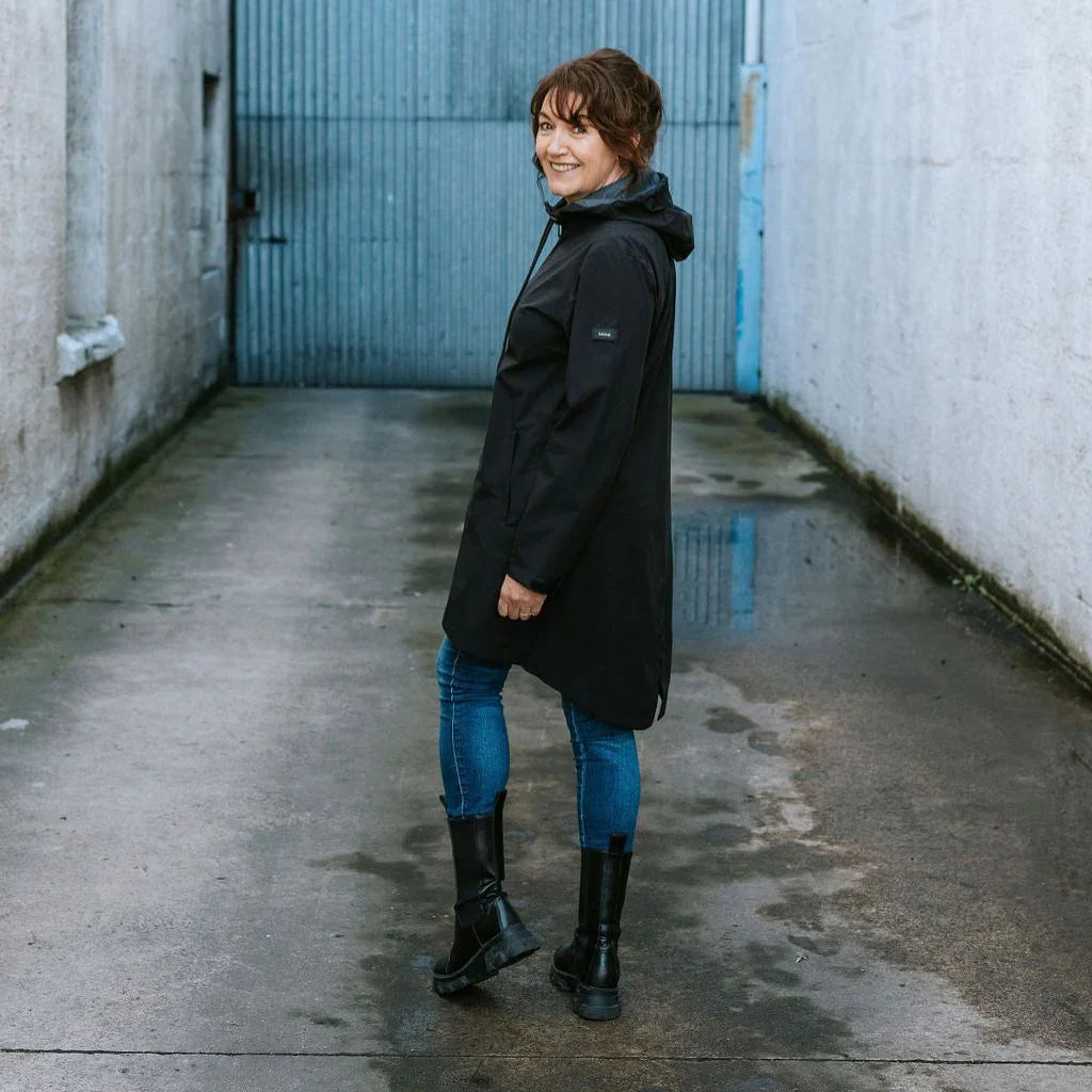 Woman wearing a black Moke rain jacket, blue jeans, and black boots in an alleyway.