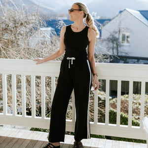 Woman wearing a Moke black sleeveless jumpsuit with white side stripes on a white balcony.