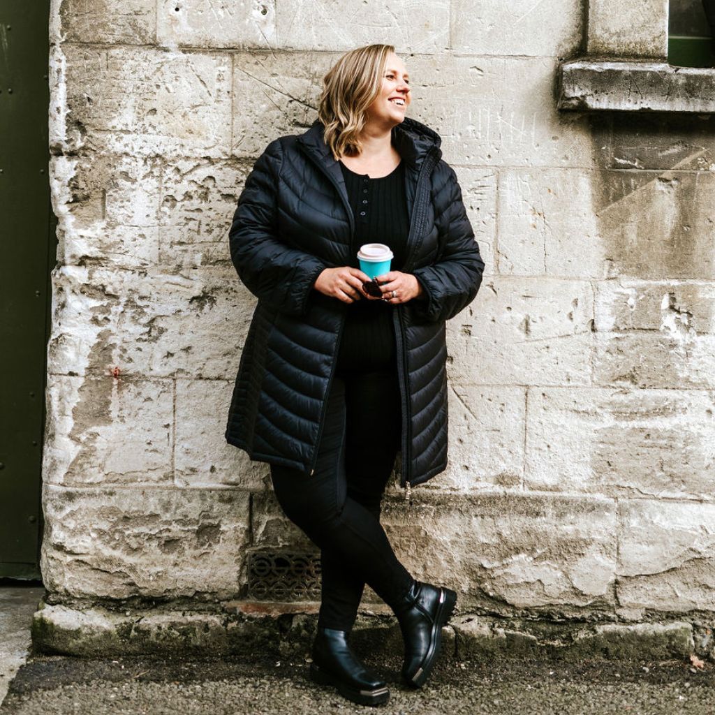 Woman in a black coat standing against a stone wall holding a blue cup.