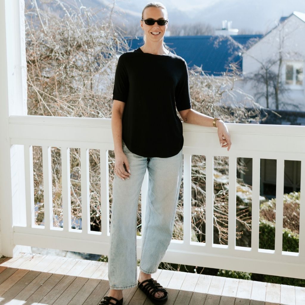 A woman wearing a Moke black t-shirt and light wash jeans standing on a white porch.