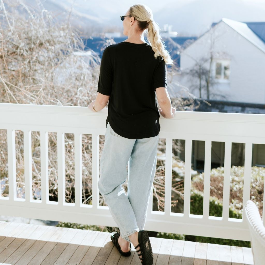 Woman wearing a black Moke t-shirt and light blue jeans standing on a white balcony.