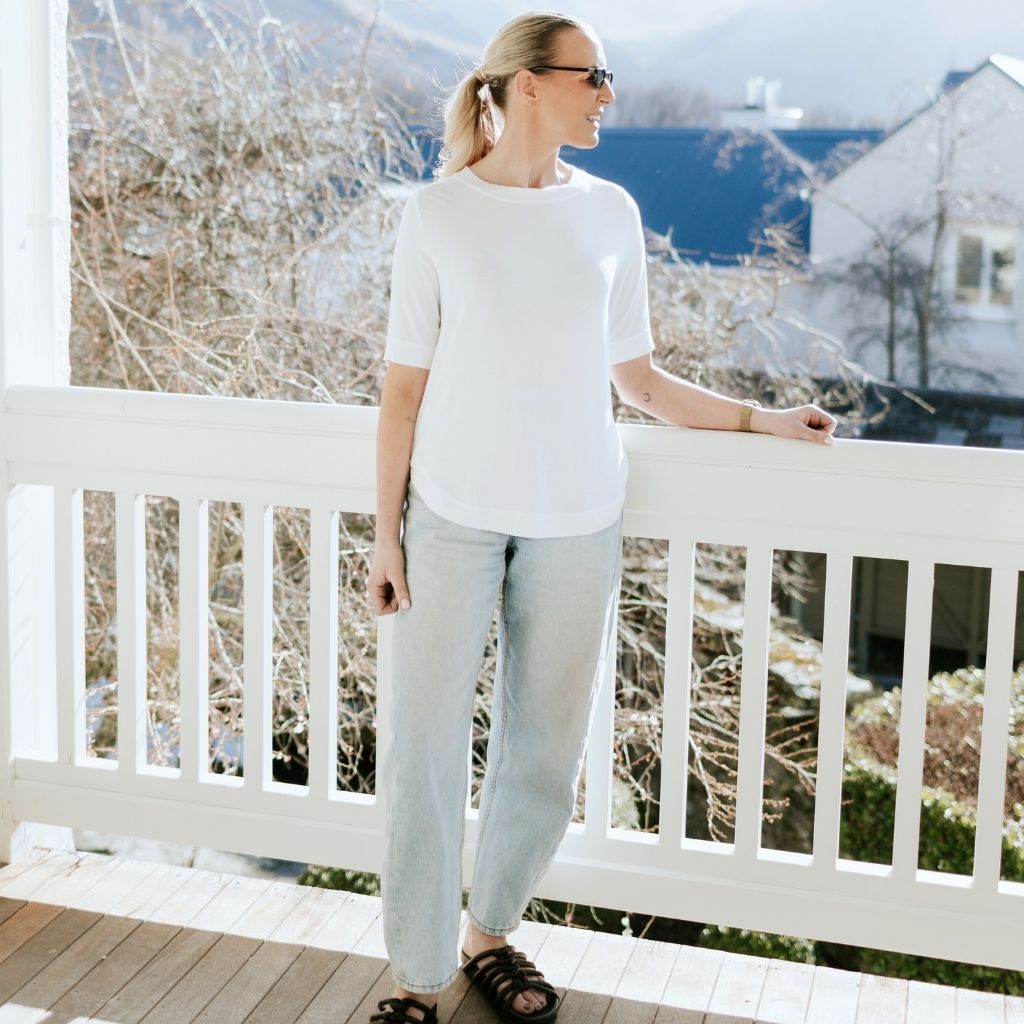 Woman wearing a white Moke t-shirt and light blue jeans standing on a white balcony.