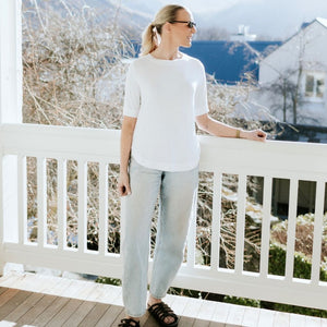 Woman wearing a white Moke t-shirt and light blue jeans standing on a white balcony.