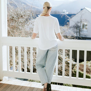 Woman in a white Moke t-shirt and light wash jeans leaning on a white balcony railing.
