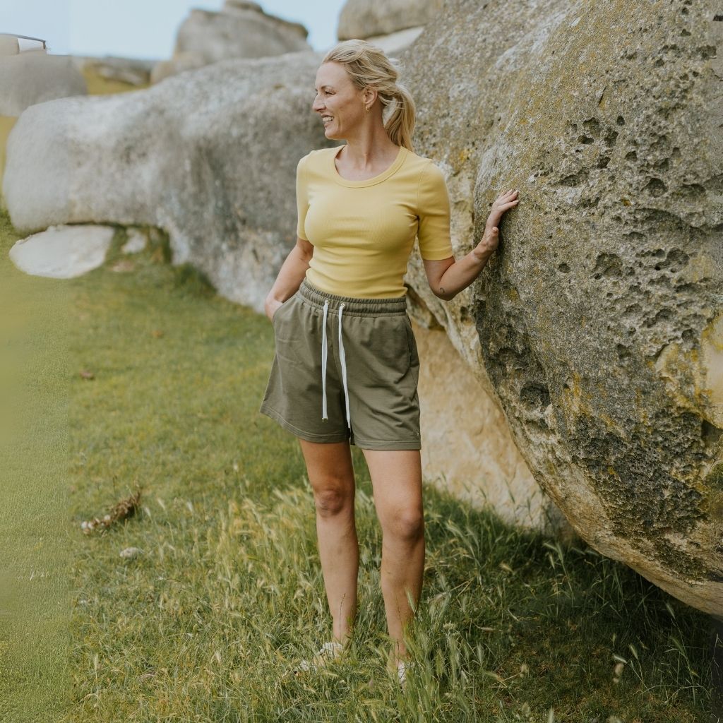 Woman in yellow shirt and green shorts standing next to large rocks in a natural setting